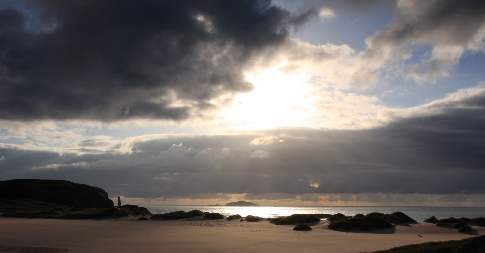 A beach at sunset with heavy clouds over the sea in the distance and pristine sand in the foreground. Hills and a sea stack are visible to the left.