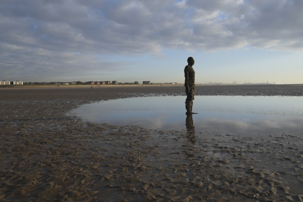 Image of a cast iron statue standing on a beach at dusk and low tide with coastal buildings on the horizon in the background