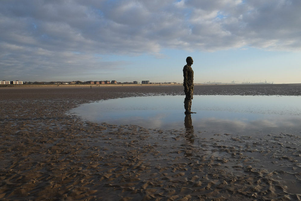 An art installation by Antony Gormley. A cast iron statue standing on a beach at dusk and low tide with coastal buildings on the horizon in the background.