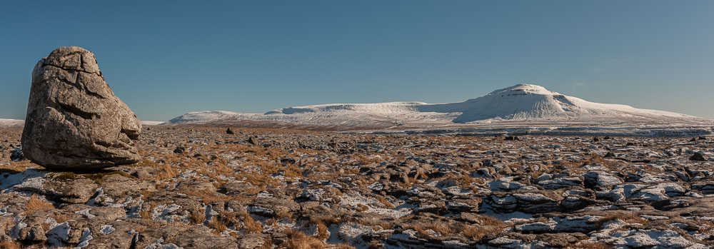 Panorama showing a large boulder on a snow flecked moor with bue sky and Ingleborough hill in the distance