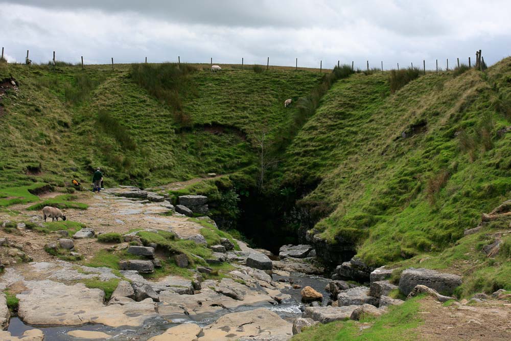 Gaping Gill shaft entrance, a river flows into a dark hole at the end of a shallow valley