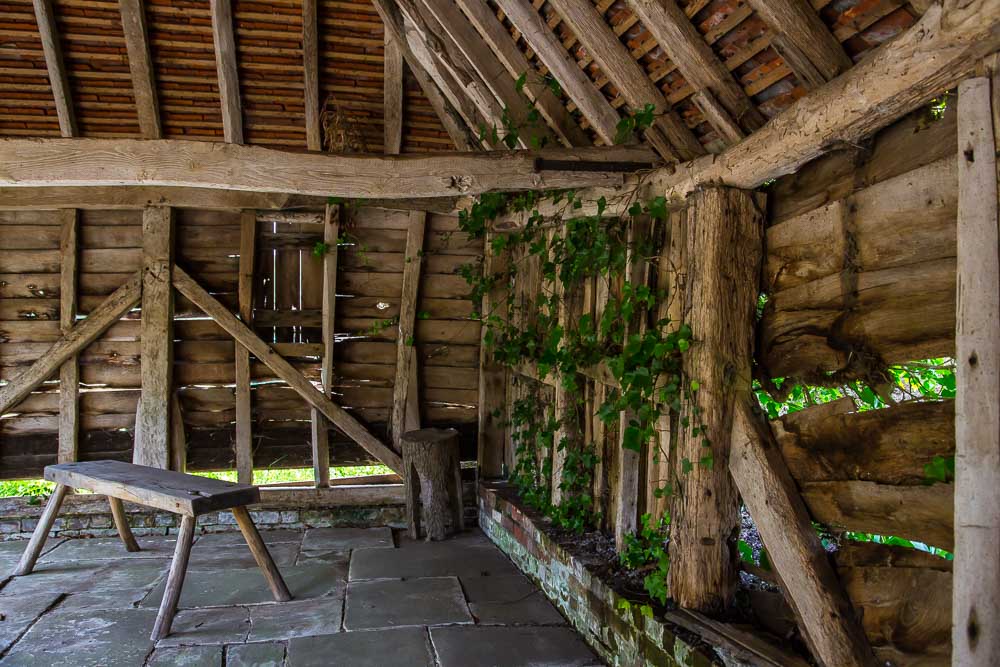 Image of the inside of a wooden barn. A simple wooden bench is visible in the corner and ivy is growing through the gaps in the old oak panelled walls.