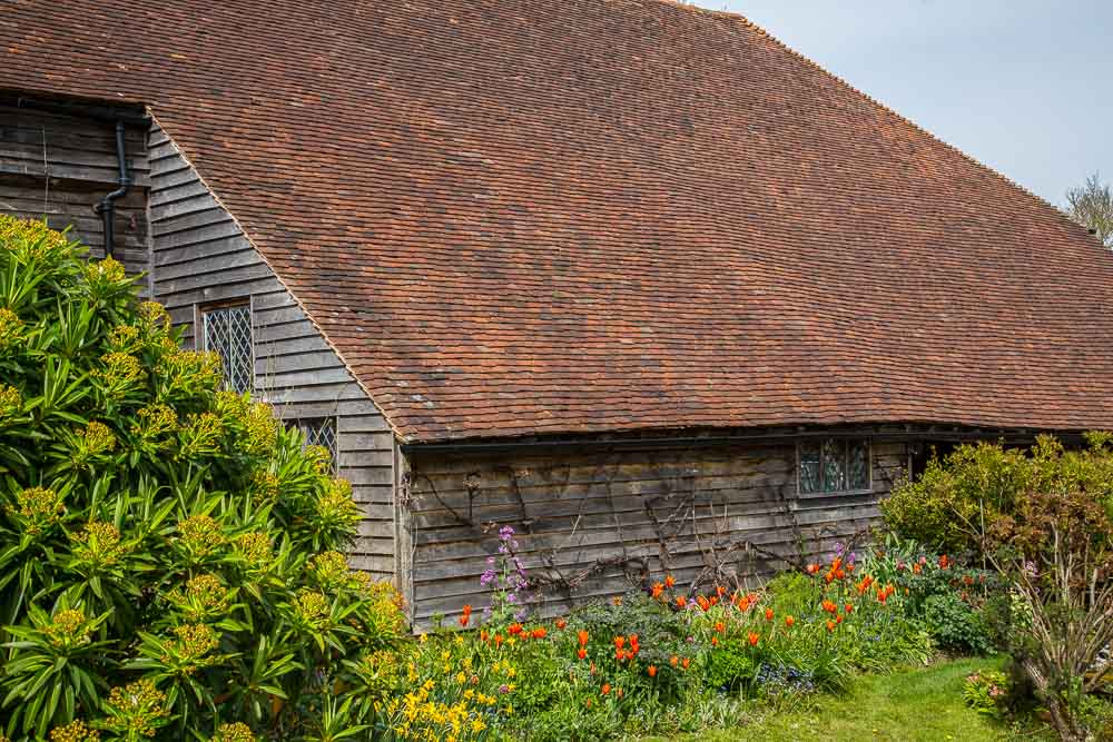 A long sloping roof made from terracotta tiles on an old wooden barn. red tulips growing along the barn wall facing the camera.