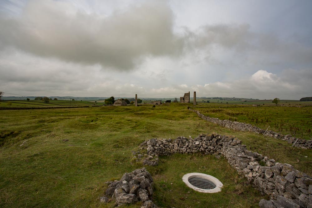 A mine shaft capped with a metal grill with derelict mine workings in the distance