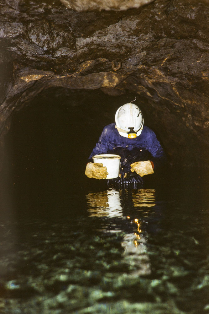 a person wearing blue overalls, a white helmet with lamp attached standing waist deep in water in a mine tunnel