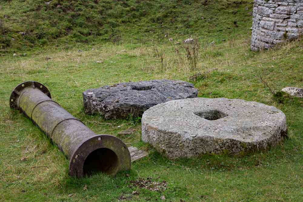 An old iron pipe and two mill wheels lying on grassy ground.