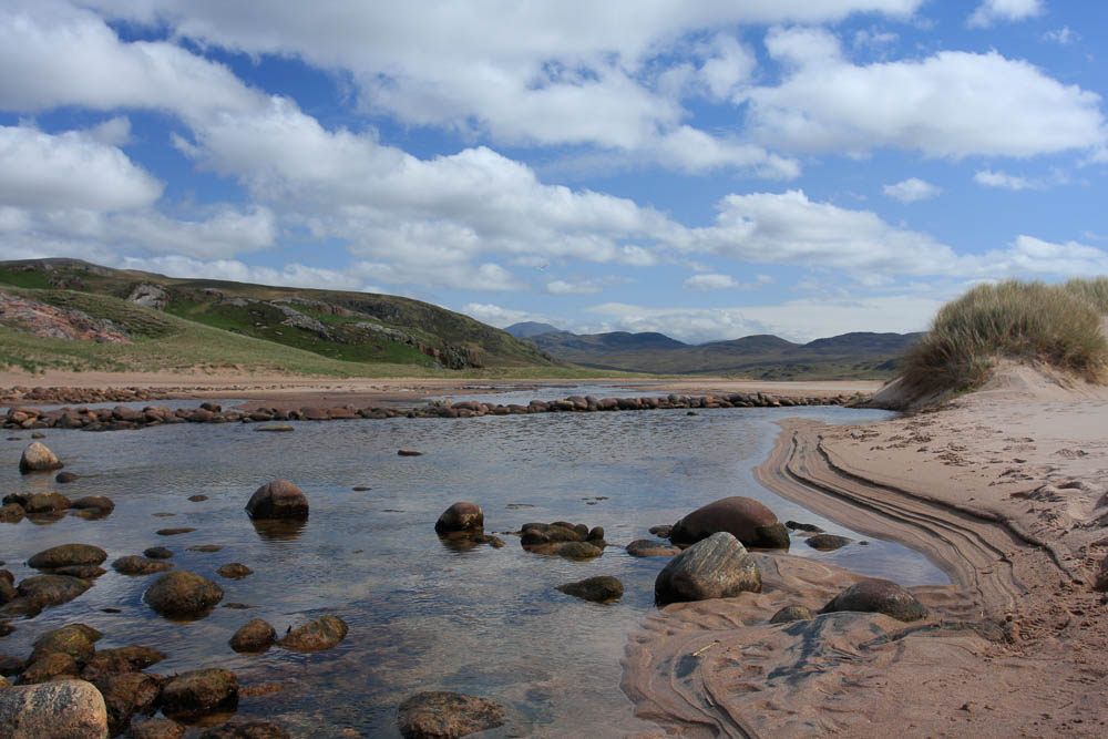 Image of a shallow river flowing from hills in the background with blue sky.