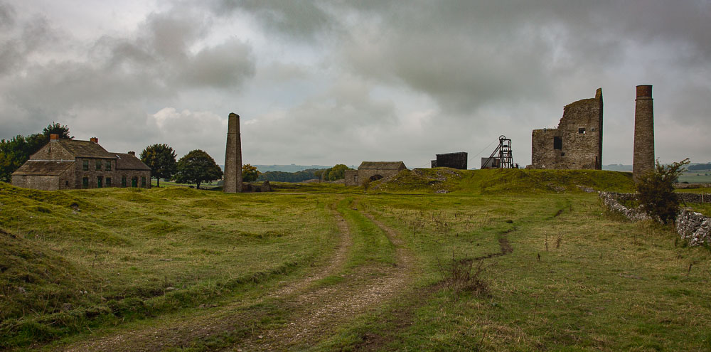 Mine workings and buildings (including house, chimneys and winding gear) with a rough gravel track leading up to them.