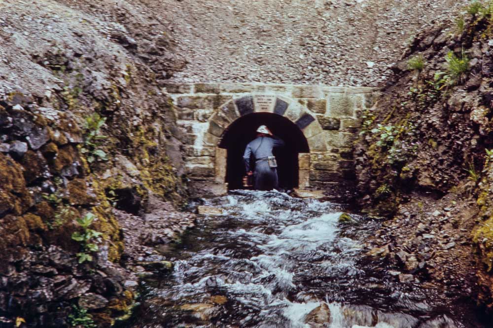 A stream flowing from a stone arch entrance and a man wearing a miner's lamp and helmet entering the tunnel.