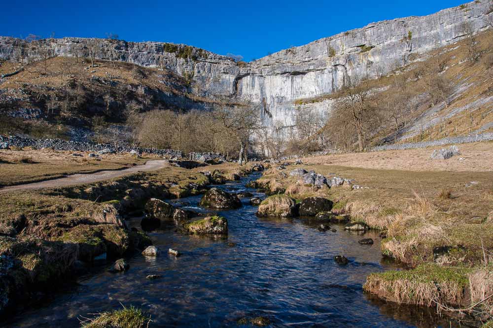 Malham Cove, a limestoone cliff face, taken from beneath with a river running towards the camera from the cliff bottom