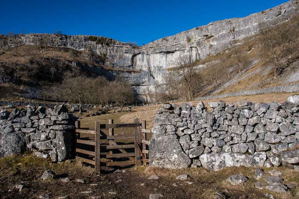 Malham Cove, a limestone cliff face, taken from the bottom with a drystone wall and wooden gate in the foreground
