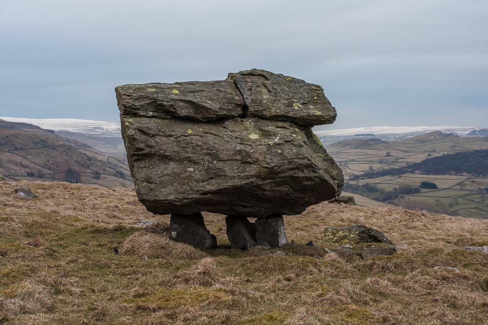 A large boulder held up by three smaller rocks, snow clad hills in the background