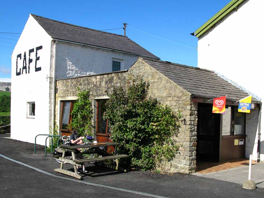 The Three Peaks Cafe, the word "cafe" is written in large black letters on the white gable end wall with a person sitting outside at a picnic table