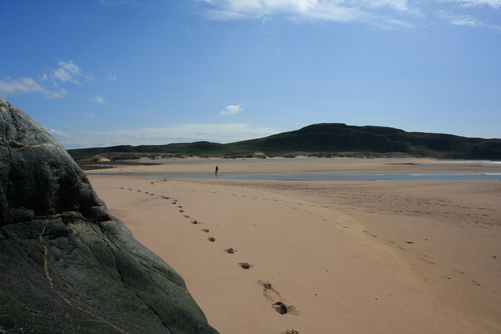 A track of fresh footprints in pristine sand leads to a person in the distance on a deserted beach.