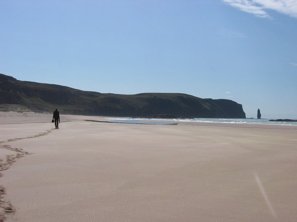A person walking along a deserted beach on a sunny day with cliffs and a sea stack in the distance beneath a blue sky.