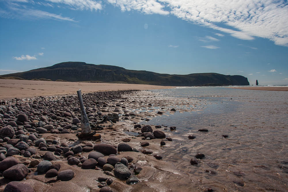 Image of a an aeroplane propellor sticking up out of the shingle on a beach on a sunny day just at the water's edge. Blue sky over hills and a sea stack are visible in the background.