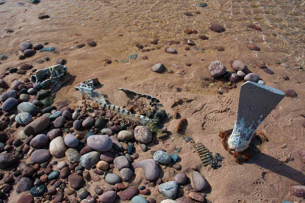 Image of the remains of an engine and propellor part buried in the sand and shingle of a beach.