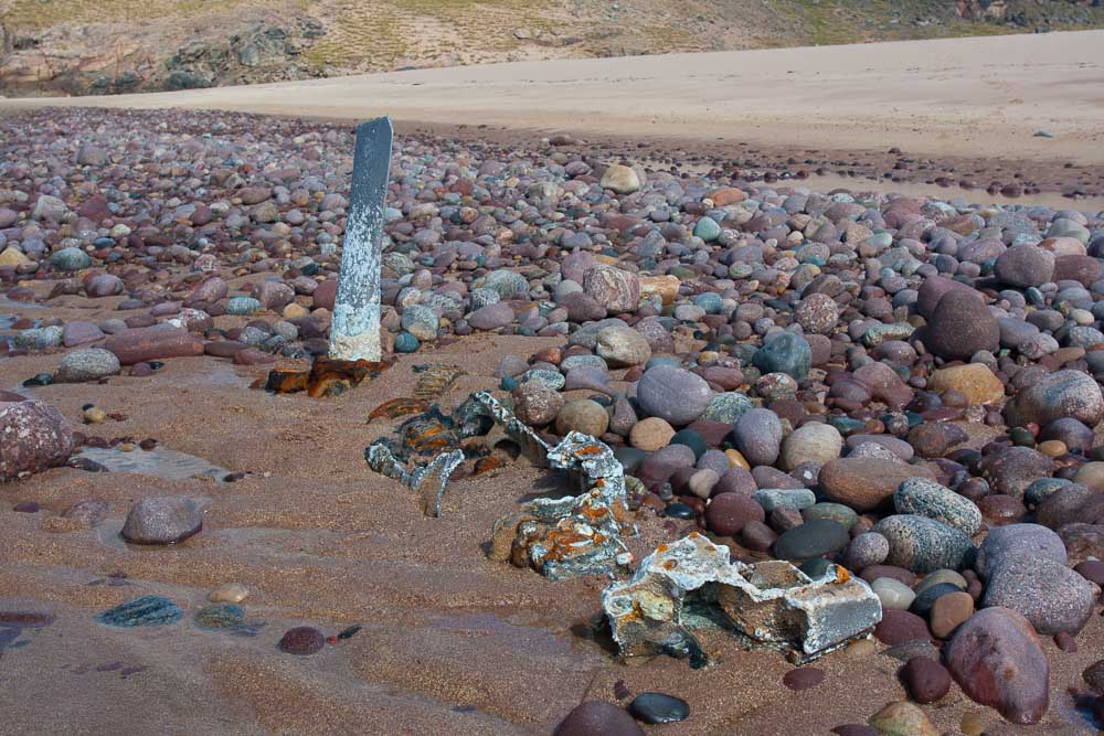 Image of the remains of an engine and propellor part buried in sand and shingle on a beach.