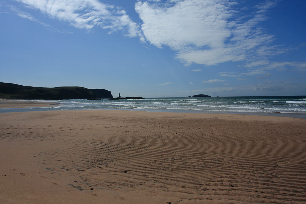 Image of a sandy beach with cliffs and a sea stack in the distance. Shallow waves are breaking on the beach beneath a blue sky.