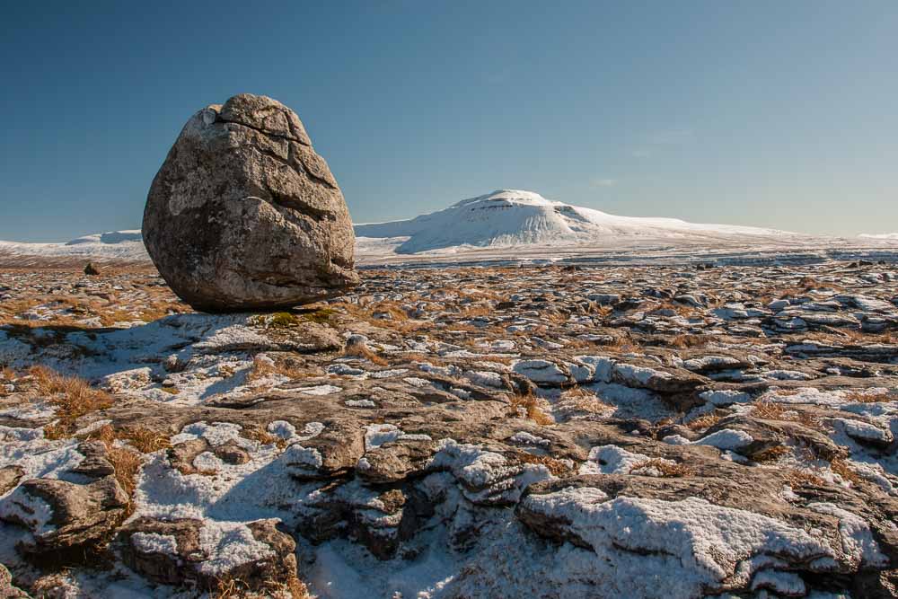 A large boulder on a moor with snow on the ground and Ingleborough hill in the distance