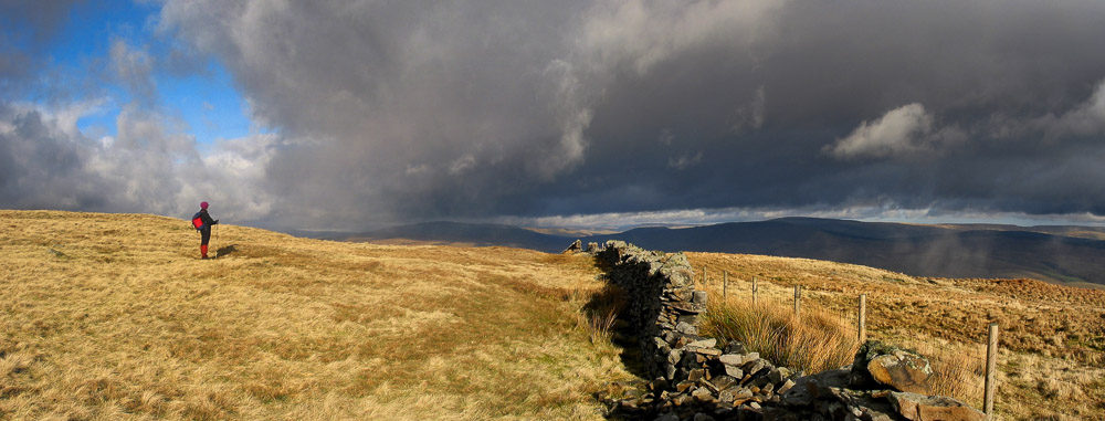 a person facing a drystone wall across a moorland with very heavy grey skies overhead