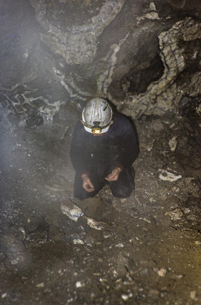 A man kneeling and examining rocks inside mine workings, crystals visible in the rocks behind.