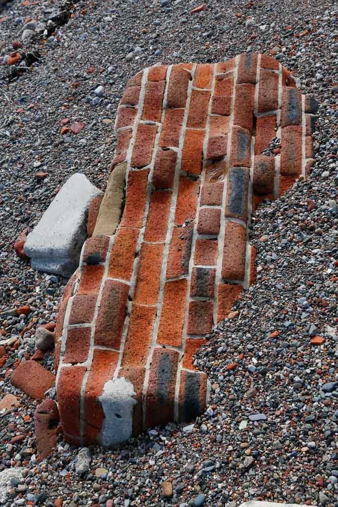 A section of brick wall lying on its side partly buried in crushed stone and brick and eroded into a wavy texture by the sea