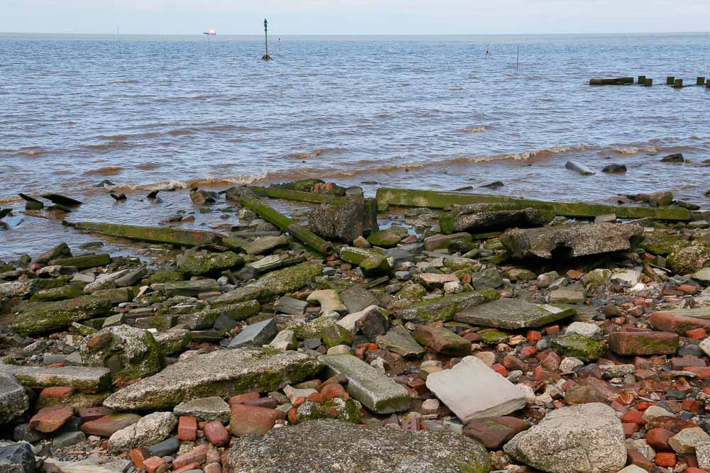 A number of discarded concrete lamp posts lying at the water's edge on a beach