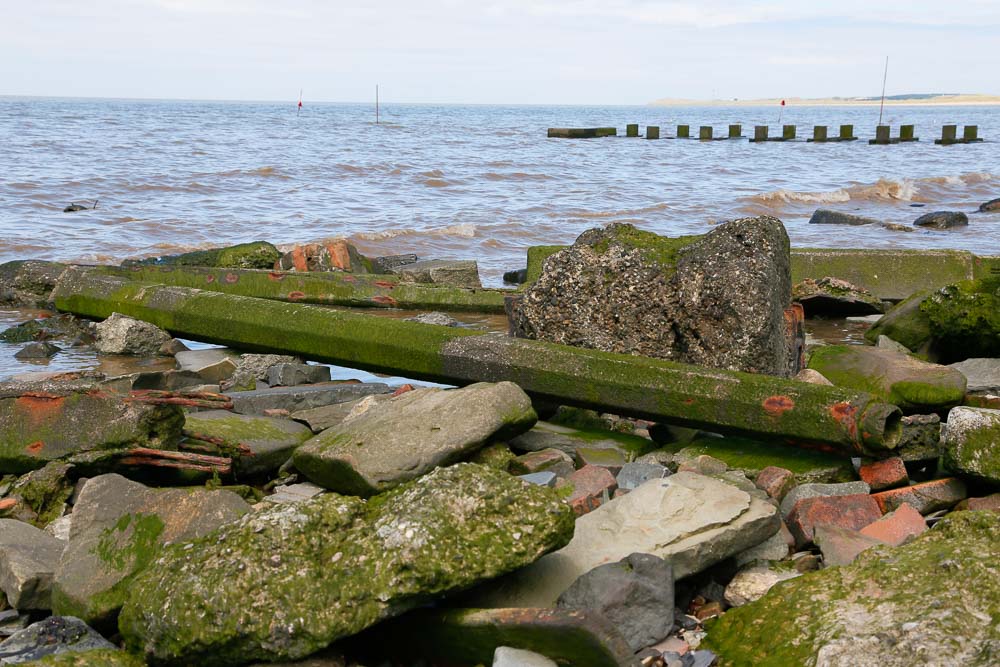 A number of discarded concrete lamp posts lying at the water's edge on a beach