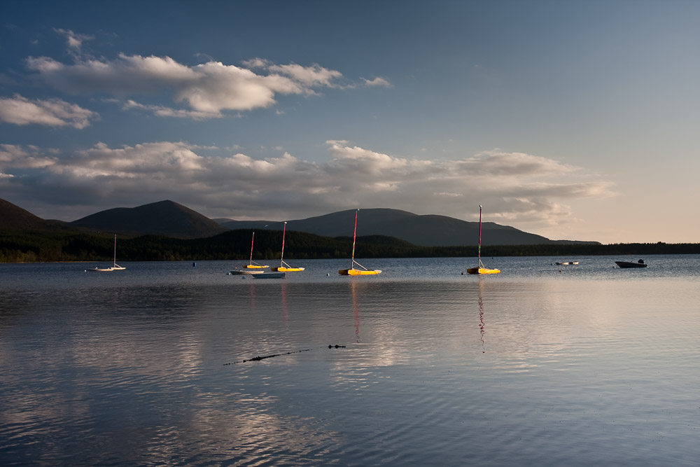 A lake in the evening light with sailing boats and dark mountains in the background