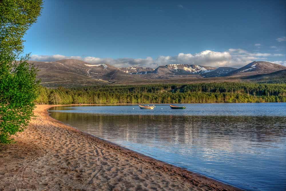 A sandy beach on a calm lake shore in the foreground with two boats visible in the mid ground and forest and mountains in the distance