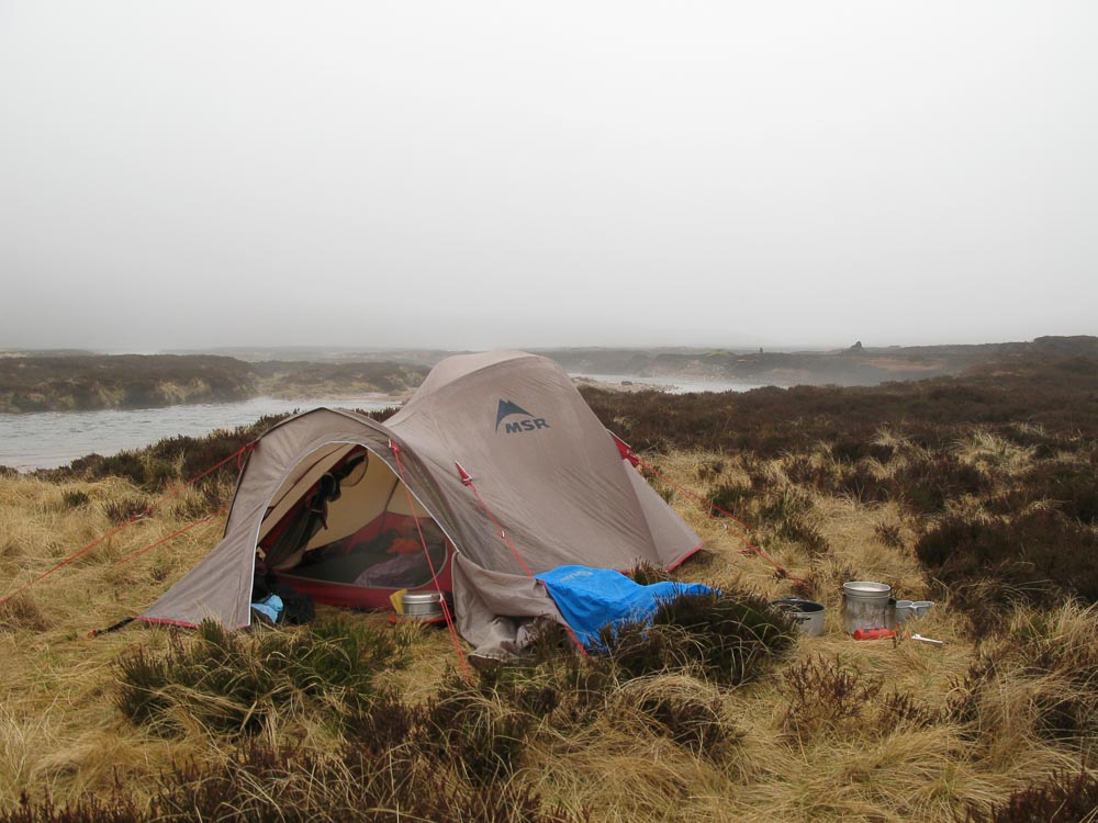 A small tent in mist and rain with cooking stove and pans on one side and a river on the other