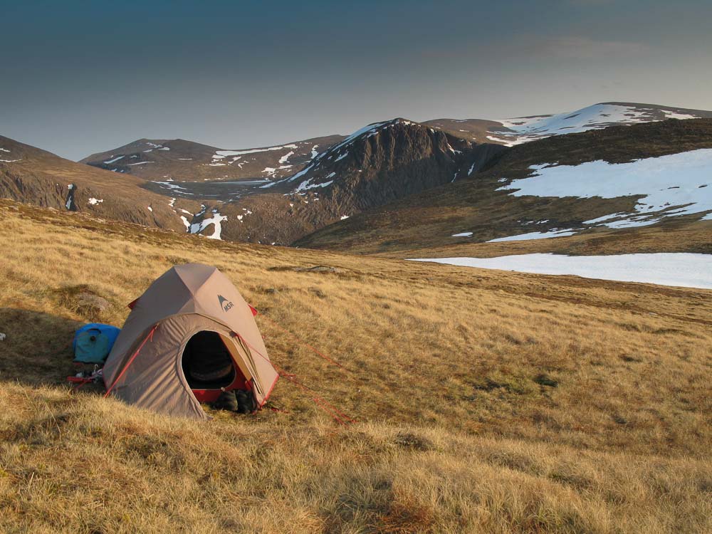 A small tent pitched on a grassy slope with snow covered hills and cliffs around it