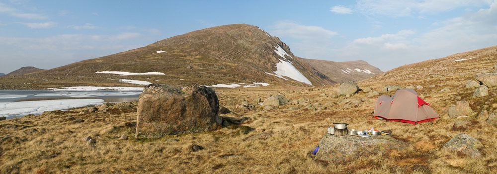 A small tent with cooking equipment outside amongst grass and boulders next to a frozen and snow covered lake